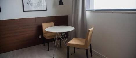 Una habitación pequeña y moderna con una mesa redonda blanca y dos sillas, una obra de arte geométrica enmarcada en la pared y una ventana con cortinas.
