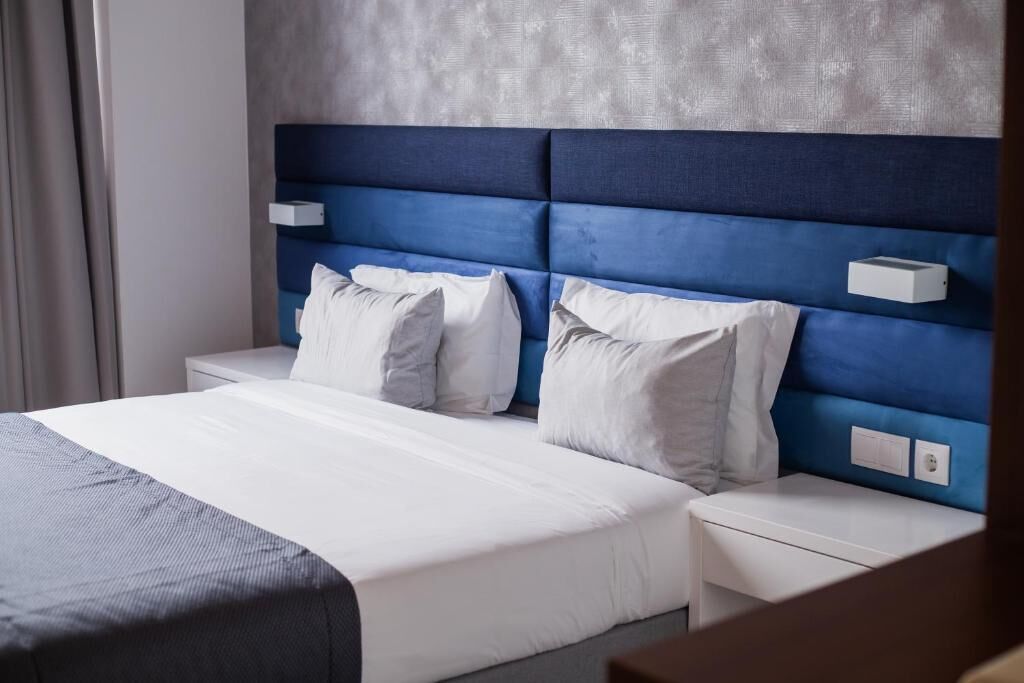 A hotel room with a large bed, white bedding, and a blue headboard. There are two bedside tables and a small nightstand.