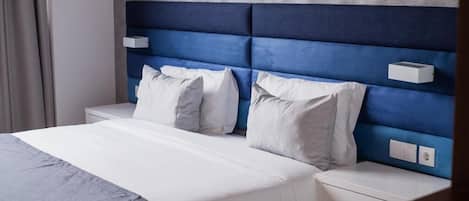 A hotel room with a large bed, white bedding, and a blue headboard. There are two bedside tables and a small nightstand.