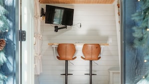 A small room with two brown leather bar stools, a mounted television, and a wooden table. - Tiny Blanco Retreat - River Tiny Home ~ Hill Country (Wimberley)