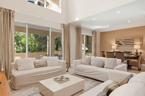 Un salón amplio con dos sofás blancos, una mesa de centro y una zona de comedor con sillas de mimbre.