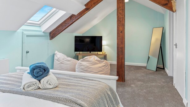 A bedroom with a sloped ceiling, wooden beams, a bed with rolled towels, a TV, and a mirror. - Large House in Central Ulverston | Sleeps 10 - Pass the Keys (Ulverston)