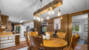 A wooden dining area with a round table and chairs, a kitchen with wooden cabinets, and a ceiling with exposed beams and hanging light fixtures. - Forest Cabin Near Sekiu Fish & Hunt Basecamp (Clallam County)