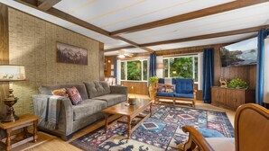 A cozy living room with a sofa, armchair, and coffee table, wooden beams on the ceiling, and a large window with curtains. - Forest Cabin Near Sekiu Fish & Hunt Basecamp (Clallam County)