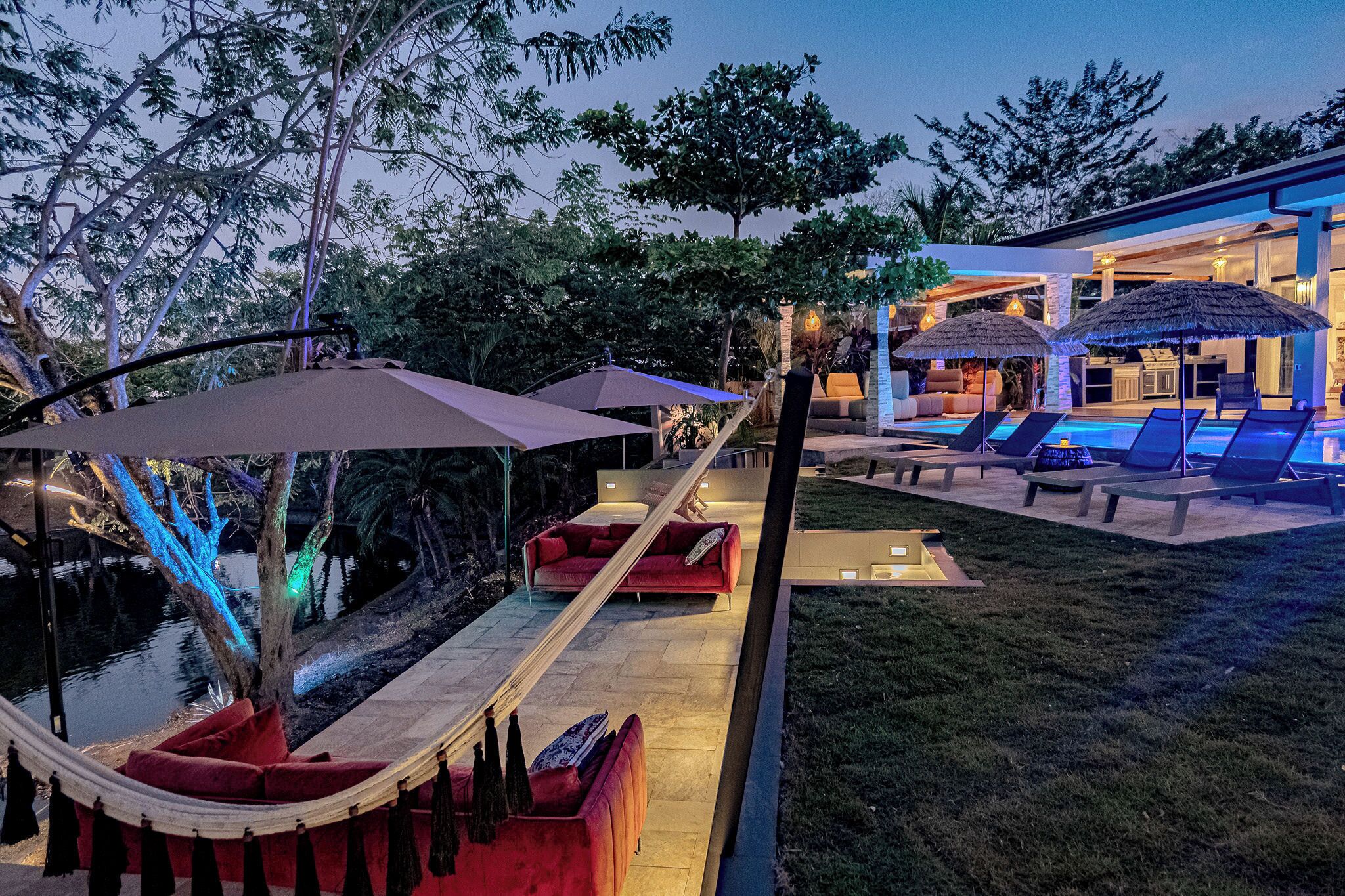 A modern outdoor lounge area with a hammock, red sofa, and lounge chairs under umbrellas, adjacent to a building with a pool and outdoor kitchen.