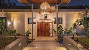 Entrada d'un edifici modern amb una porta de fusta, una gran làmpada penjant i un jardí ben cuidat.