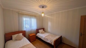 A bedroom with two single beds, a nightstand, a ceiling light fixture, a window with sheer curtains, and a wooden door. - Patio do garajau (Caniço)