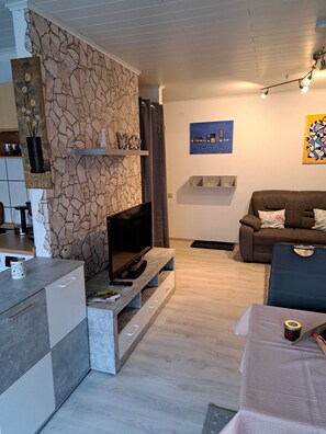 A modern living room with a stone accent wall, a flat-screen TV, a brown leather sofa, and a dining area with a table and chairs. - Cozy 2-room ground-floor apartment with wheelchair access and a terrace   (Ludwigswinkel)