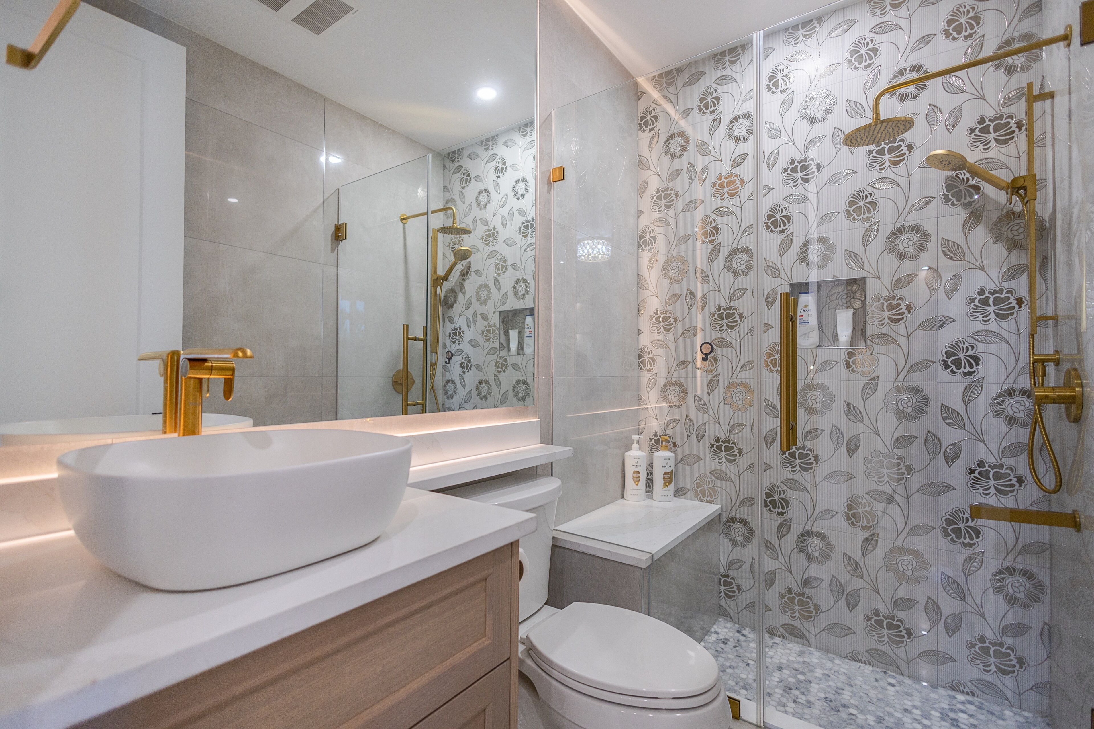 A modern bathroom with a large white sink, gold fixtures, a toilet, and a shower with floral wallpaper.