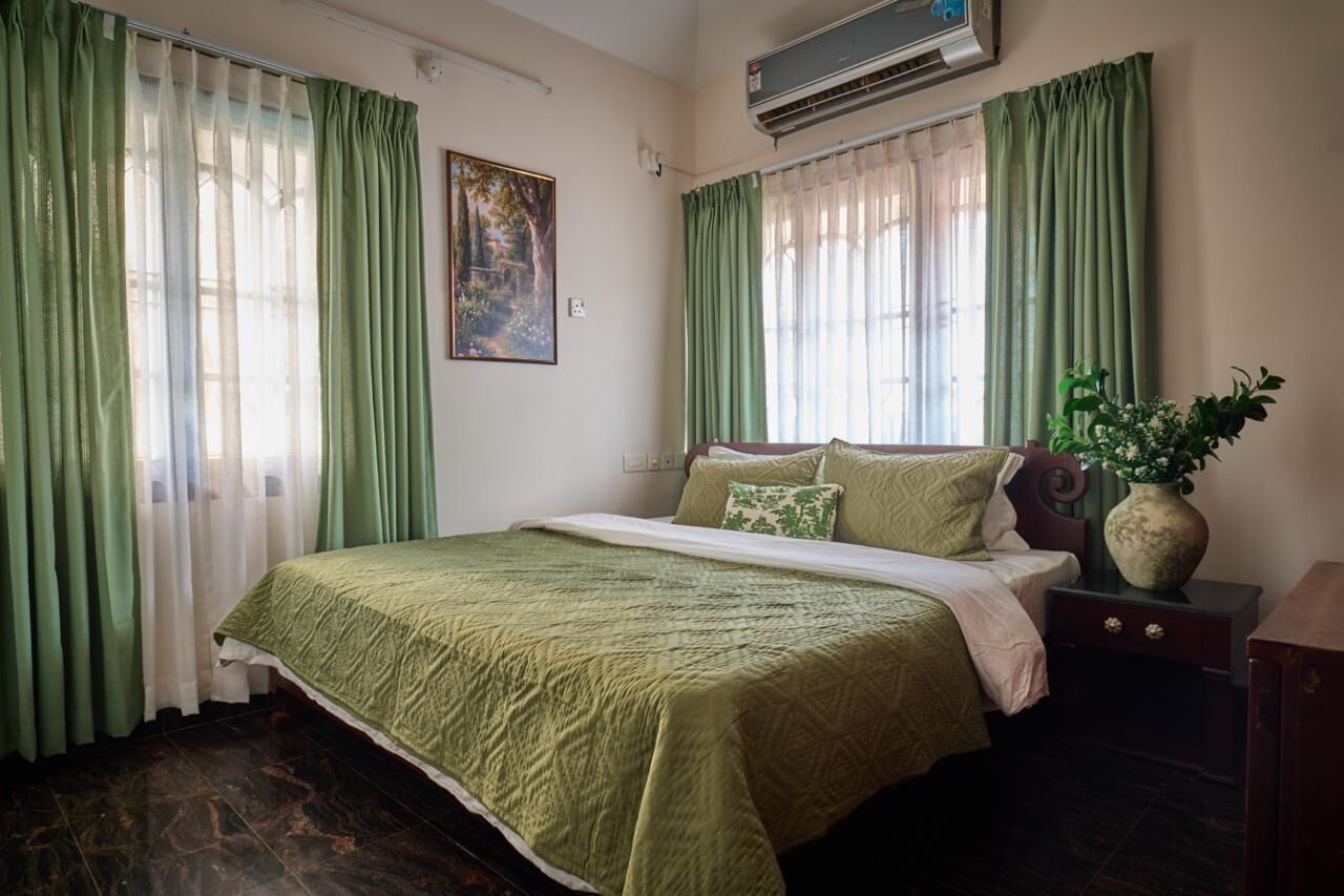 Une chambre à coucher bien rangée, avec un lit, des rideaux verts, un climatiseur fixé au mur et une petite table de chevet sur laquelle se trouve un vase.