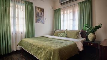 Une chambre à coucher bien rangée, avec un lit, des rideaux verts, un climatiseur fixé au mur et une petite table de chevet sur laquelle se trouve un vase.