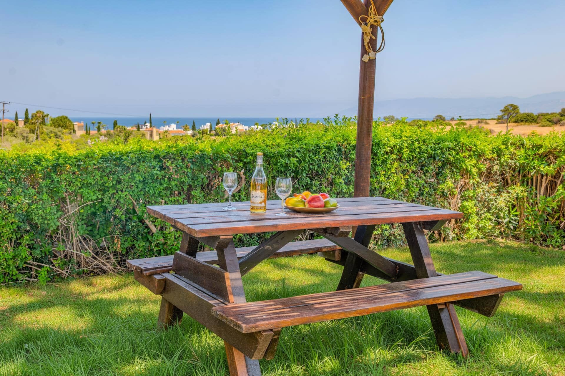 A wooden picnic table with a bottle and glasses, surrounded by greenery and a view of the sea.