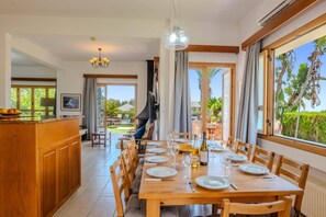 A dining area with a long wooden table set for a meal, a kitchen with wooden cabinets, and a view of a garden through large windows. - Villa Akamas Bayview by Villa Plus (Latchi)