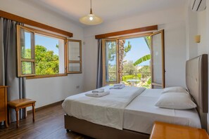 A hotel room with a bed, a small table, a chair, and a window with a view of greenery. - Villa Akamas Bayview by Villa Plus (Latchi)