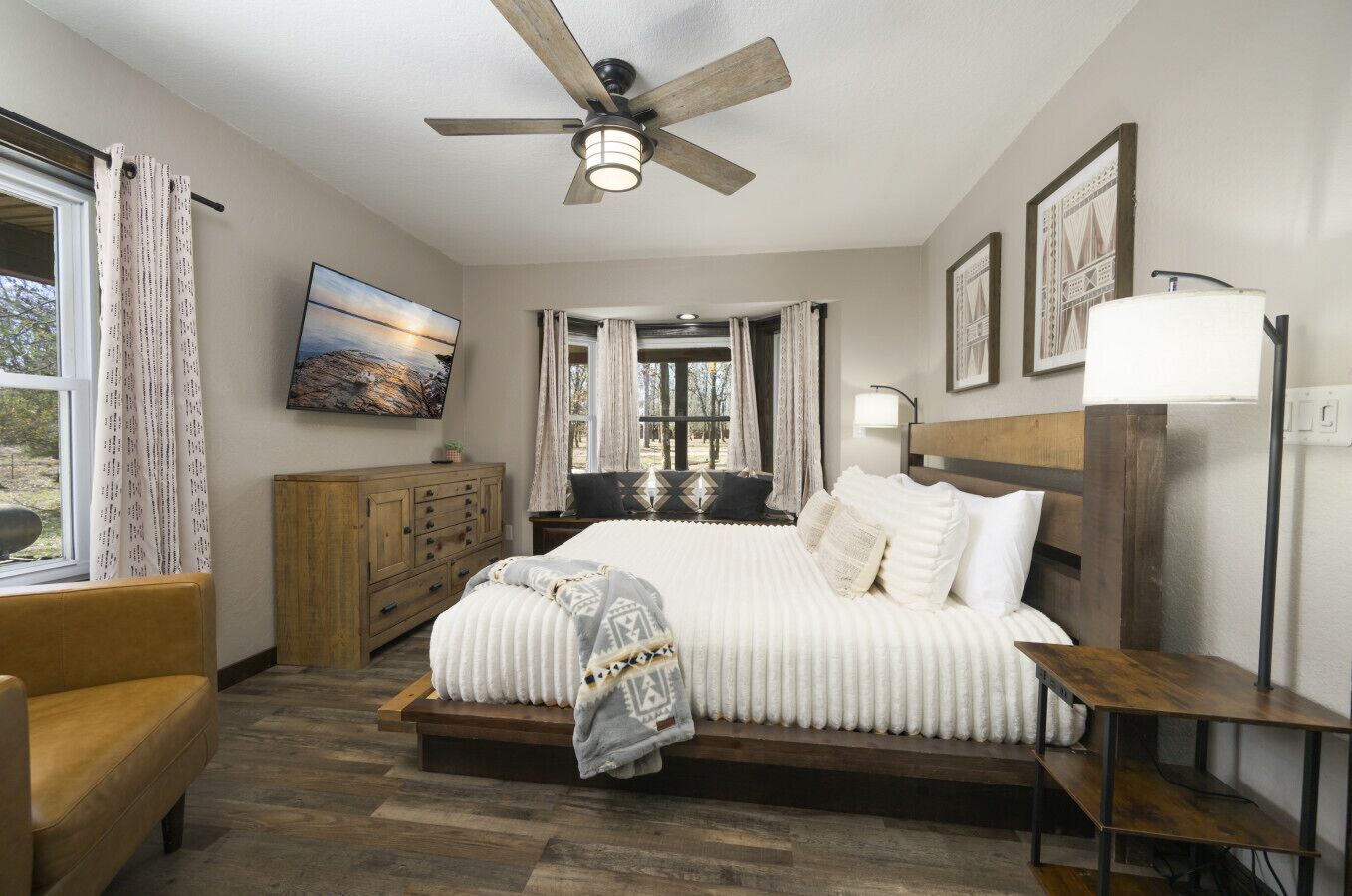 A bedroom with a bed, a wooden headboard, a nightstand, a desk, a chair, a ceiling fan, and a window with curtains.