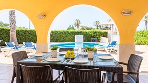 An outdoor dining area with a pool, covered by arches, and surrounded by lounge chairs and greenery. - Villa Marte by Villa Plus (Cap d'Artrutx)