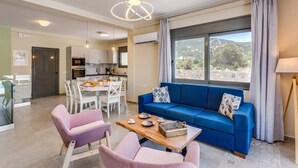 Een moderne woonkamer met een blauwe bank, twee roze fauteuils, een houten salontafel en een groot raam met uitzicht op het landschap.