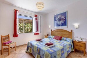 Ein Schlafzimmer mit einem Bett, einem Sessel, einem Nachttisch, einer Wandleuchte und einem Fenster mit Vorhängen.