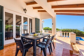 A covered patio with a dining table and chairs, a pool, and a view of the sea. - Villa S'Escaleta by Villa Plus (Punta Prima)