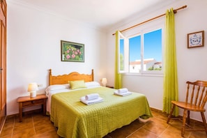 A hotel room with a bed, two bedside tables, a chair, a window with curtains, and a painting on the wall. - Villa S'Escaleta by Villa Plus (Punta Prima)