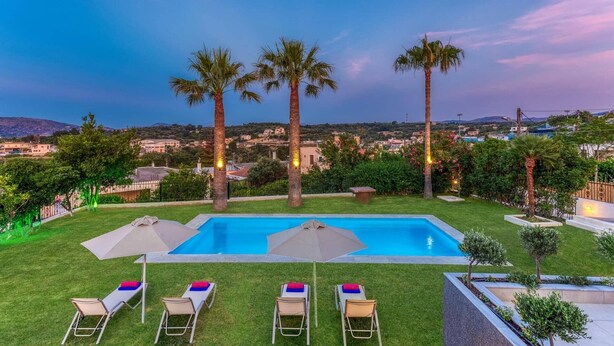 A pool area with four lounge chairs and two umbrellas, surrounded by palm trees and a view of a town in the background. - Villa Kostas Maria by Villa Plus (Episkopi)