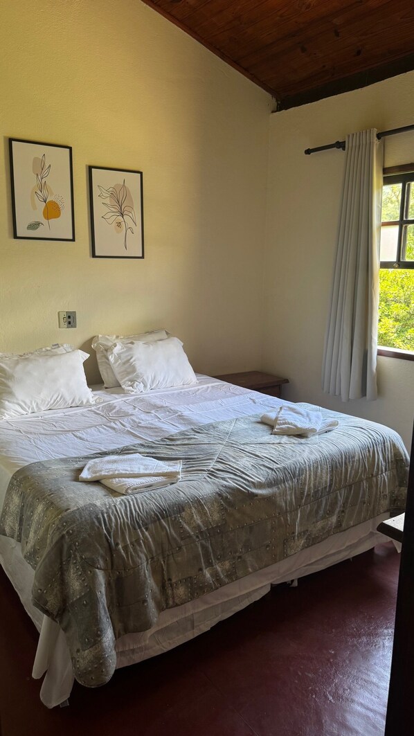 A neatly made bed with white linens and a grey patterned bedspread, a small bedside table, and two framed botanical prints on the wall. - Pousada das Cavernas (Iporanga)