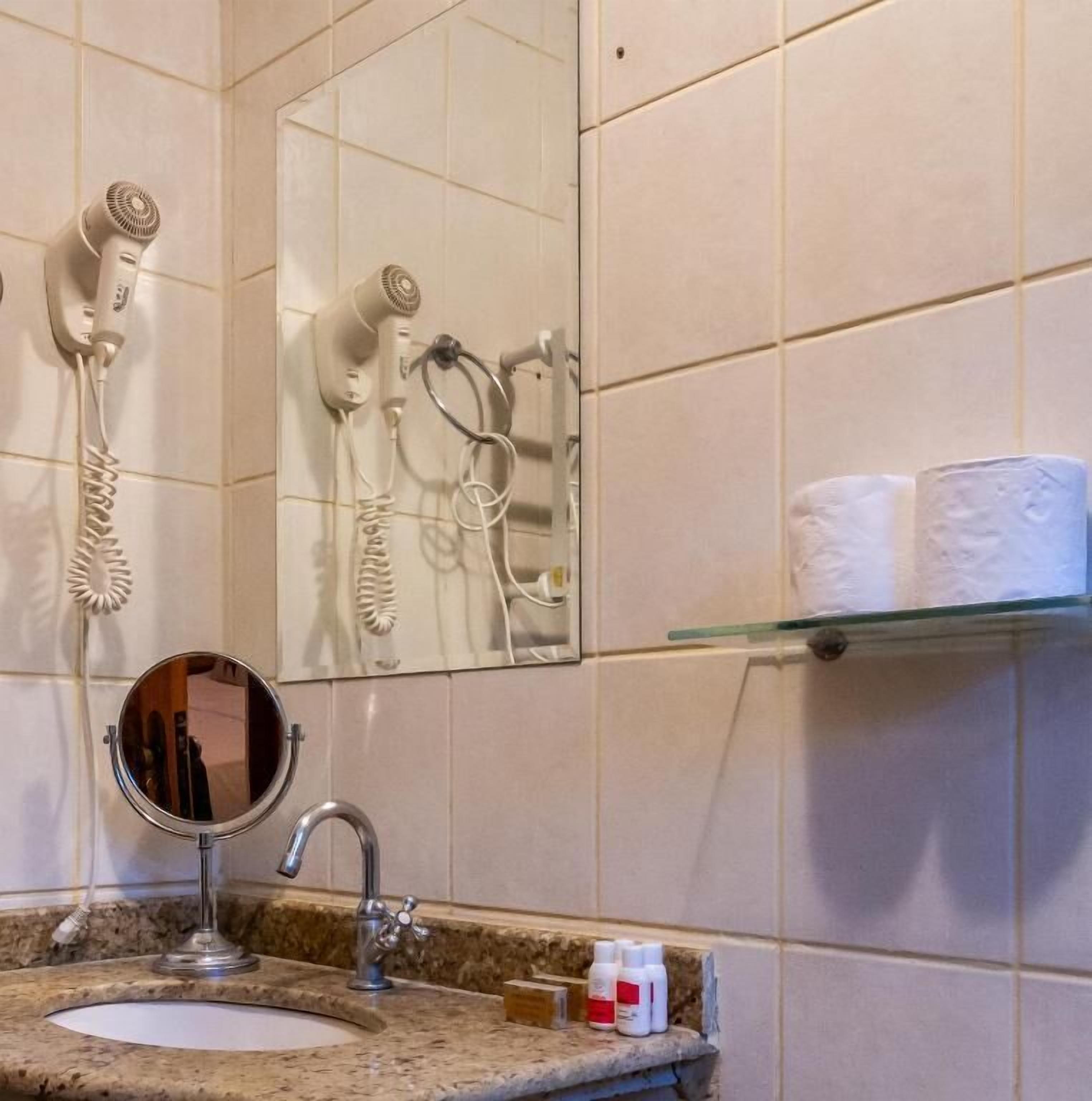 A bathroom with a sink, mirror, hair dryer, and toilet paper on a shelf.