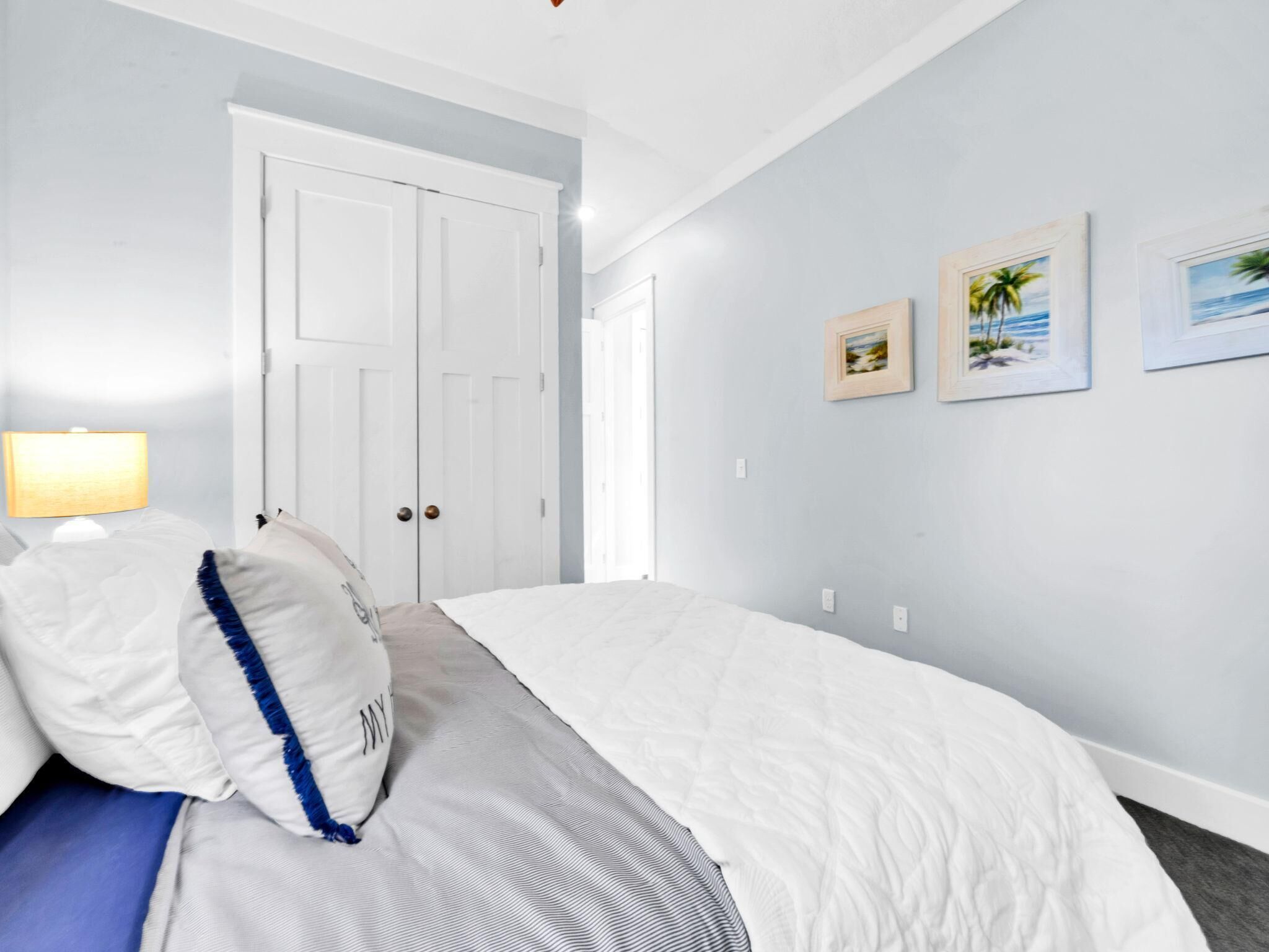 A bedroom with a bed, a nightstand, a lamp, and a door. There are three framed pictures on the wall.