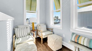 A cozy seating area with two chairs and a bench, a small table with a lamp, and a view through large windows with shutters. - The Grand Castle (Panama City Beach)