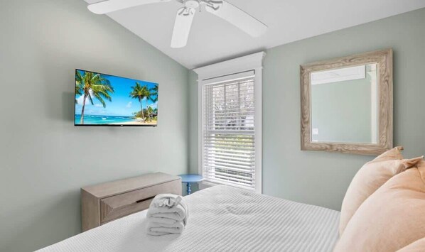 Un dormitorio con cama, televisor, ventilador de techo, ventana con persianas y un espejo enmarcado.