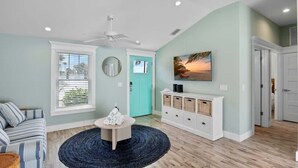 Una sala de estar con sofá, una mesa de centro, ventilador de techo, puerta con vistas y televisor montado en la pared.