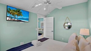Une chambre à coucher comprenant un lit, un téléviseur, un ventilateur de plafond, un miroir rond et une porte donnant sur une autre pièce.