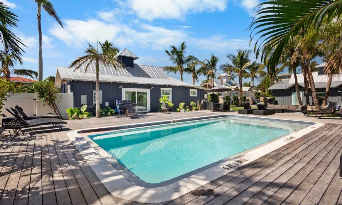A rectangular swimming pool with a wooden deck surrounding it, palm trees, and a building with a metal roof in the background.