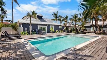 A rectangular swimming pool with a wooden deck surrounding it, palm trees, and a building with a metal roof in the background.