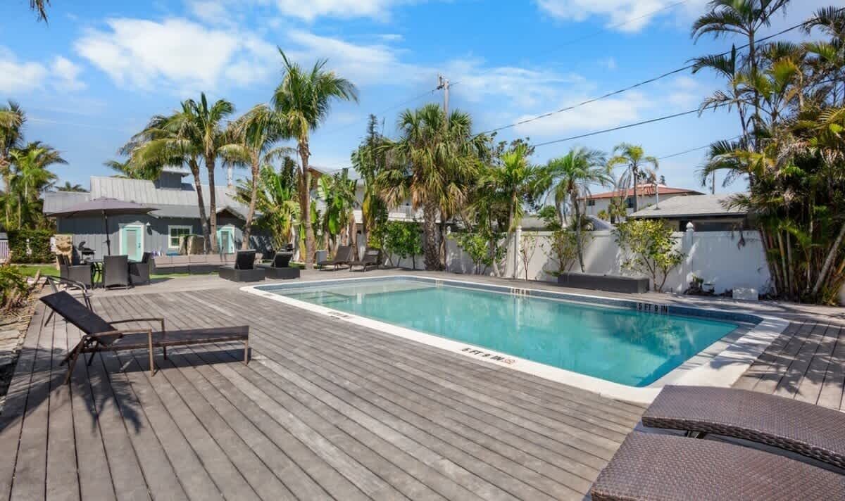 A swimming pool with a wooden deck, lounge chairs, and palm trees.