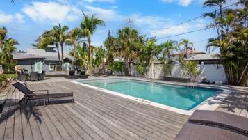 A swimming pool with a wooden deck, lounge chairs, and palm trees.