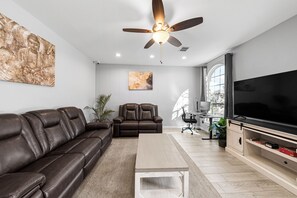 Ein modernes Wohnzimmer mit einer Ledersofa, einem Relaxsessel, einem Couchtisch, einem Flachbildfernseher und einem Deckenventilator.