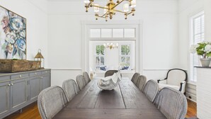 Ein Esszimmer mit einem langen Holztisch, Korbstühlen, einer Kandelaber und einem großen Fenster mit weißen Fensterläden.
