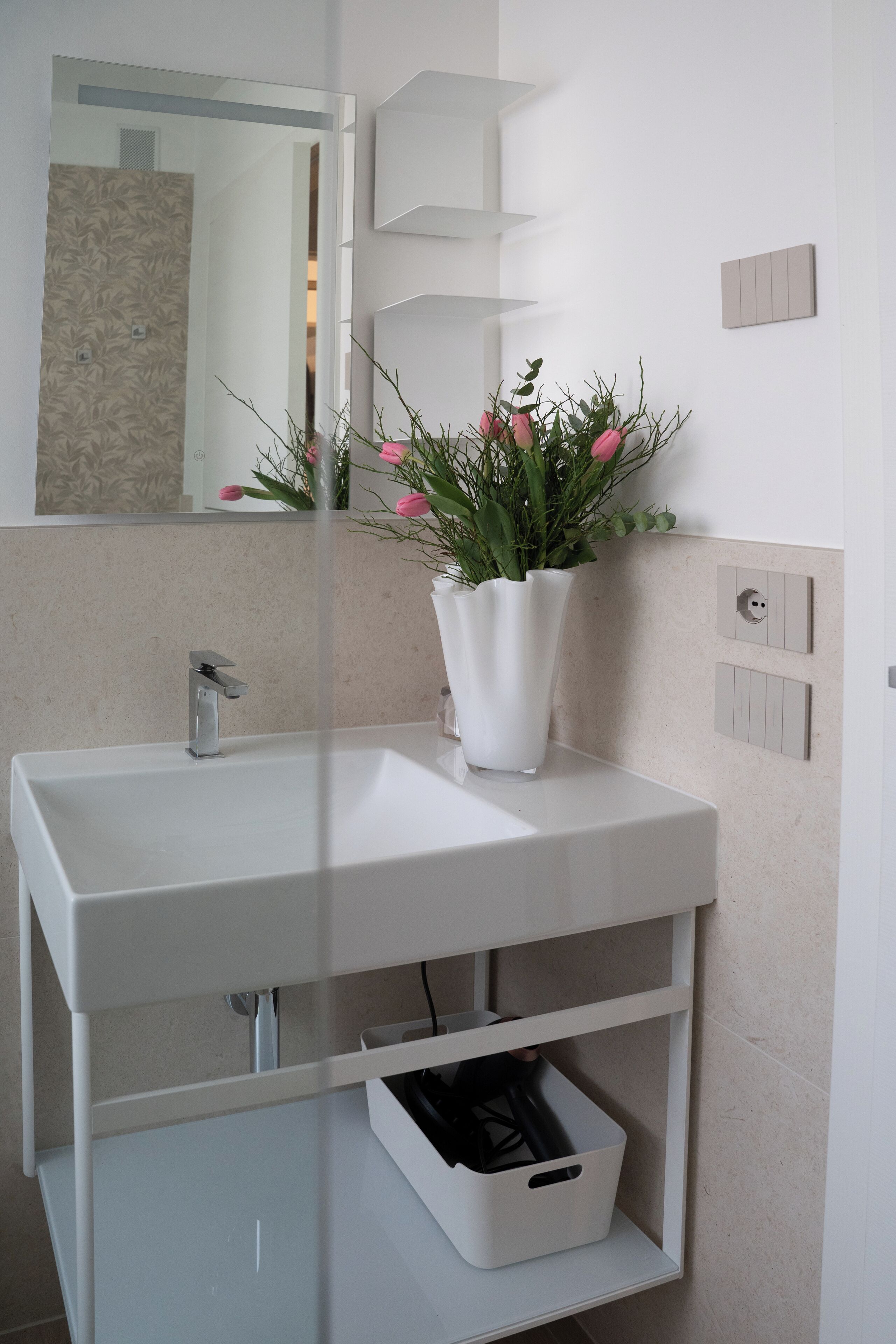 A bathroom with a white sink, a mirror, a vase with flowers, and a white cabinet below the sink.