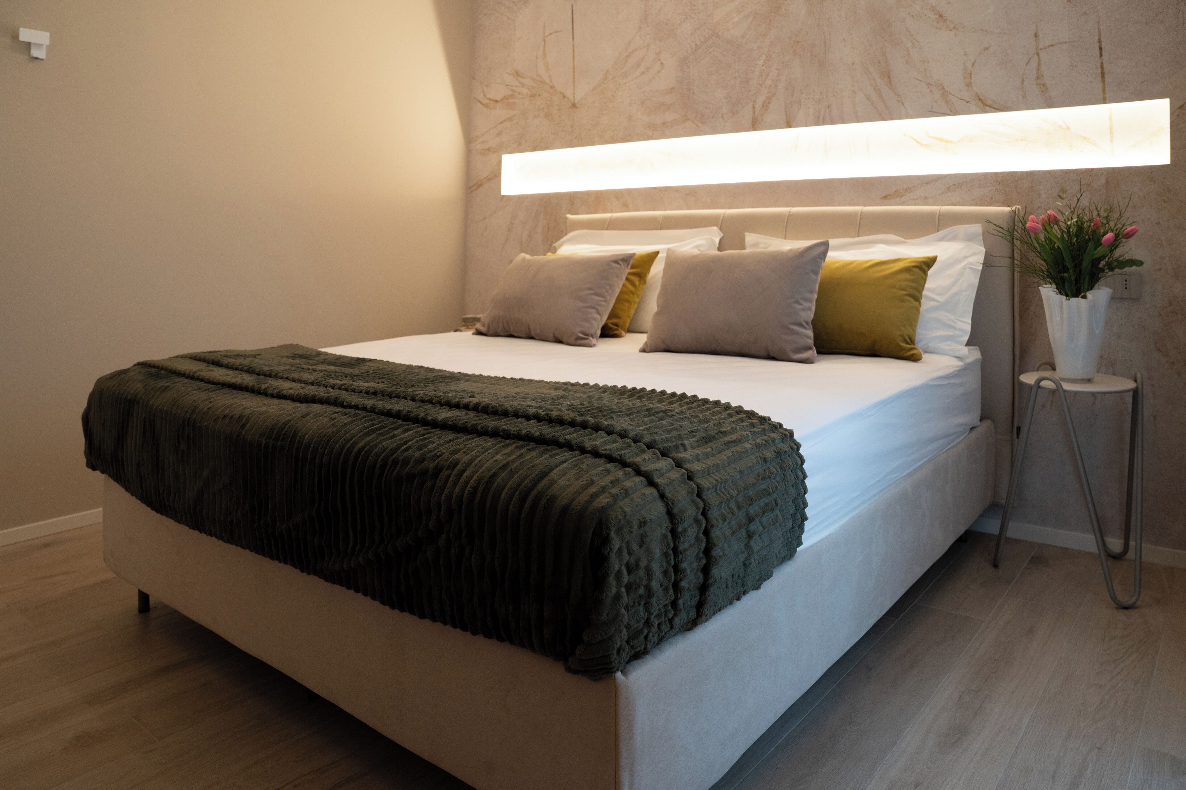 Une chambre moderne avec un grand lit, une table de chevet agrémentée d’un vase de fleurs et une tête de lit dotée d’un éclairage intégré.