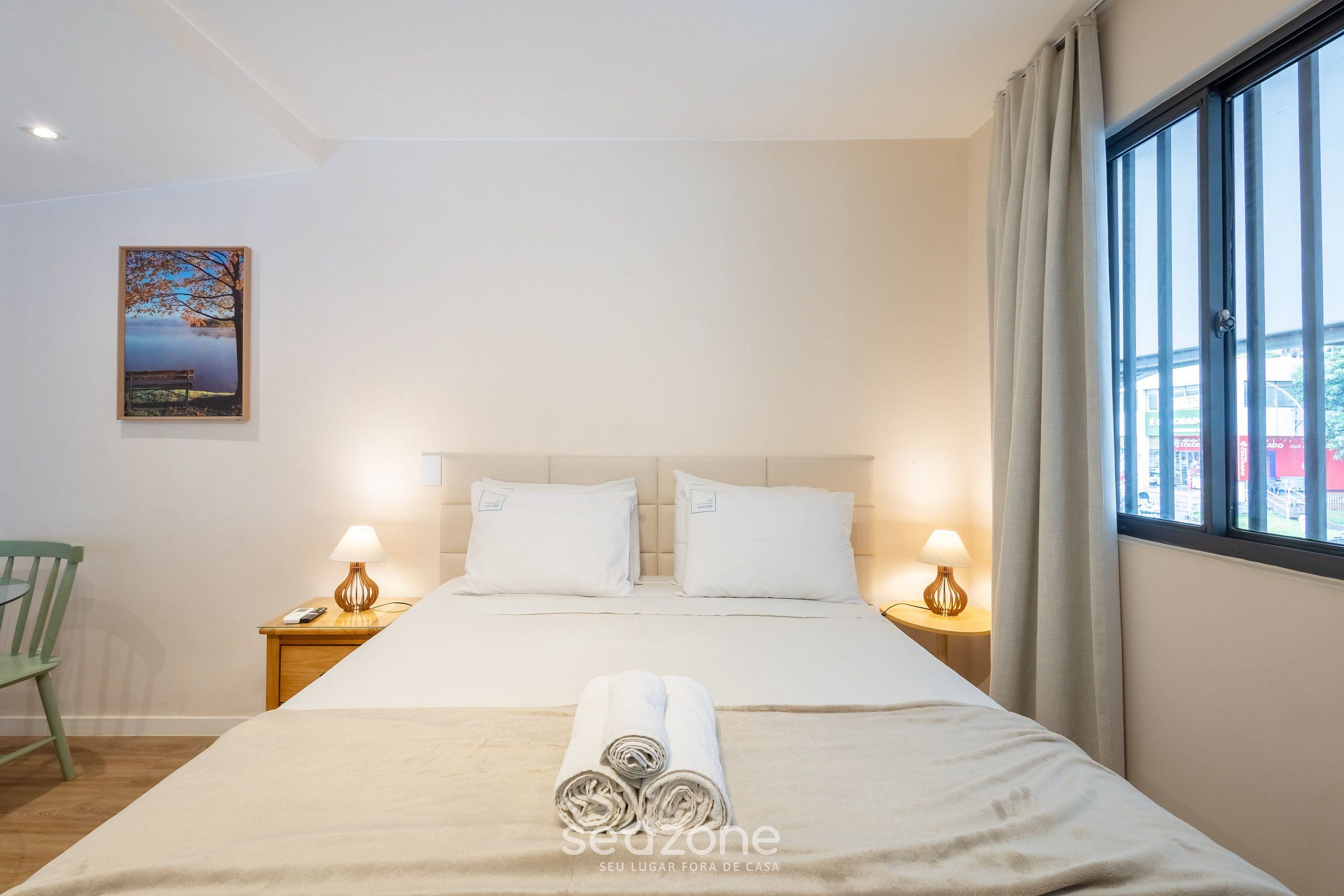 A hotel room with a bed, two bedside tables with lamps, a chair, a painting on the wall, and a window with curtains.