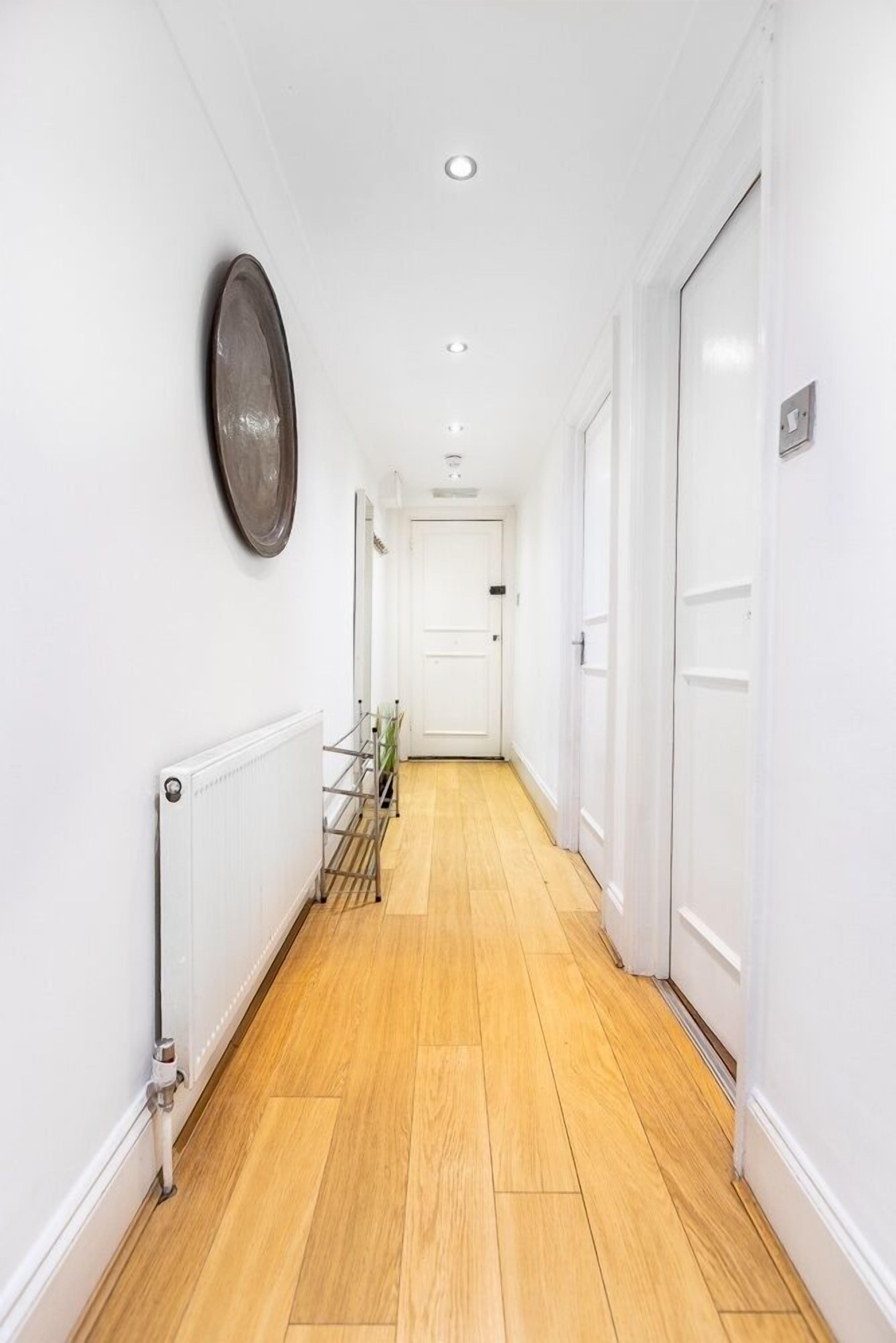 Un couloir étroit avec un plancher en bois, des murs blancs et un radiateur sur le côté gauche.
