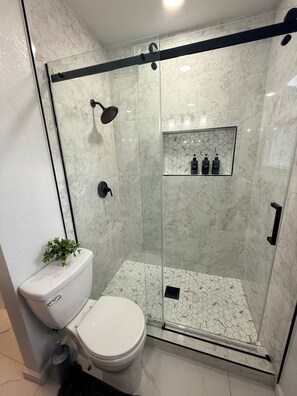 A modern bathroom with a glass-enclosed shower, a toilet, and a small shelf with toiletries. - Relaxing Cozy Back House with Backyard (Upland)
