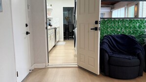 Een geopende deur die leidt naar een keuken met een houten vloer, een eethoek met een tafel en stoelen, en een donkerblauwe stoel op de voorgrond.