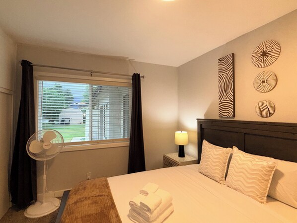 A hotel room with a bed, a bedside lamp, a wall clock, a window with curtains, and a standing fan.