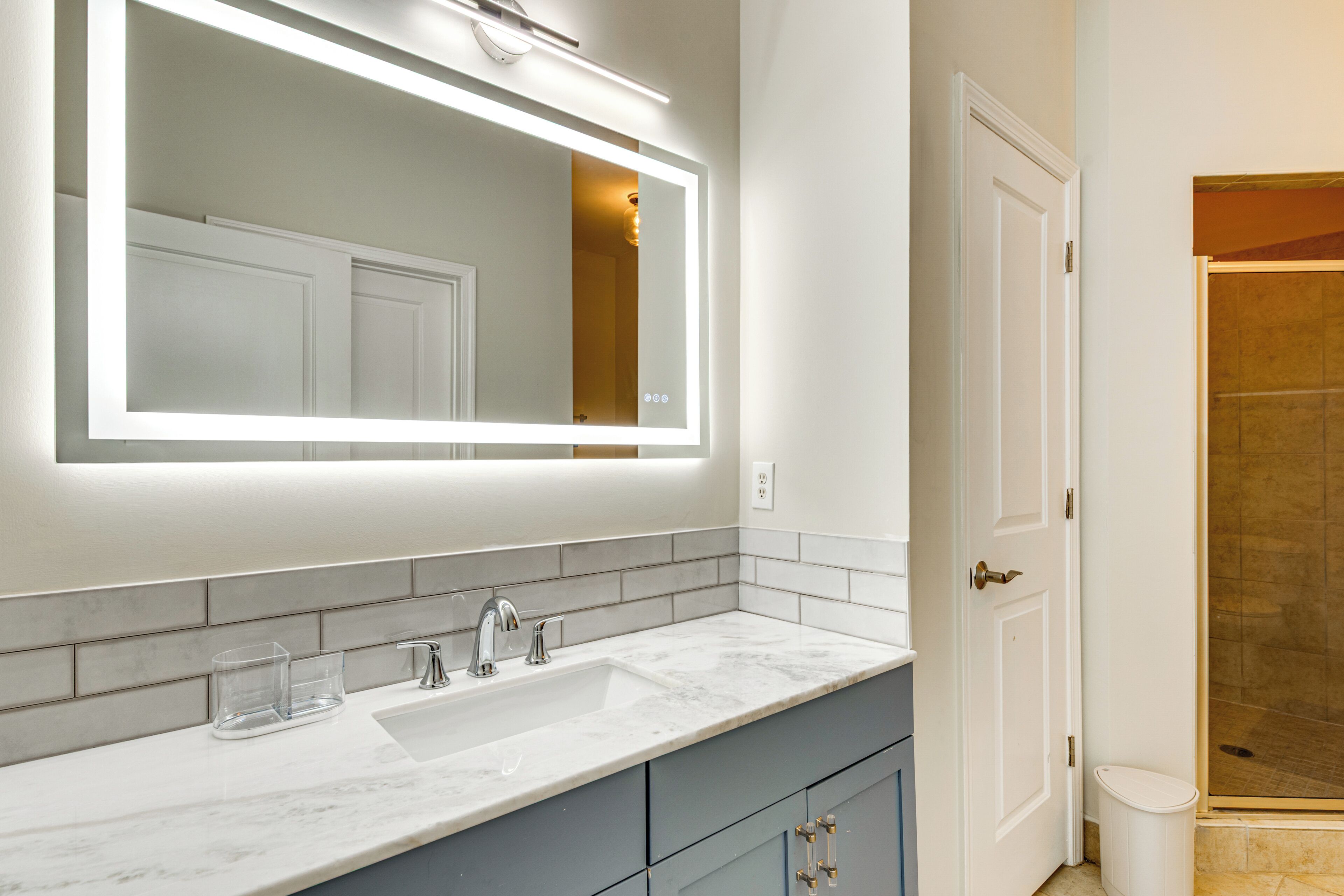 A bathroom with a white vanity, a large mirror with under-cabinet lighting, a sink, and a shower area.