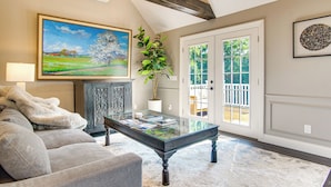 A living room with a sofa, a glass coffee table, a painting, and a door leading outside. - Summer Beach Pass: Wooded Martha's Vineyard Escape (Vineyard Haven)