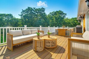 A wooden deck with a white railing, a sofa, two round tables with plants, and a grill station. - Summer Beach Pass: Wooded Martha's Vineyard Escape (Vineyard Haven)