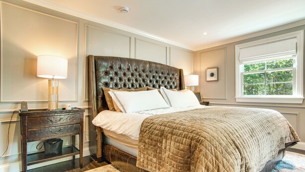 A bedroom with a large bed, a nightstand, a lamp, a window with a view of trees, and a patterned rug. - Summer Beach Pass: Wooded Martha's Vineyard Escape (Vineyard Haven)