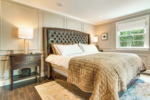 A bedroom with a large bed, a nightstand, a lamp, a window with a view of trees, and a patterned rug. - Summer Beach Pass: Wooded Martha's Vineyard Escape (Vineyard Haven)